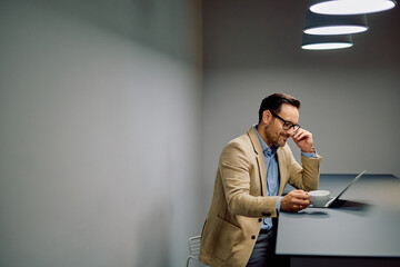 Businessman smiling while drinking coffee and working on a laptop in a modern office, taking a short break
