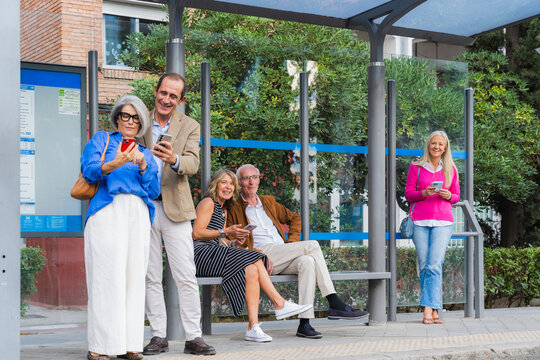 Mature friends using smartphones at bus stop waiting for public transport