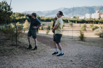 Fototapeta premium Two men with backpacks stand on a gravel path in an open park with mountains in the distance. One looks ahead while the other shields his eyes, sharing a quiet moment before hiking.