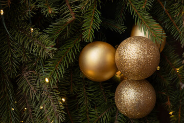 Golden Christmas ornaments hanging on fir tree branches with yellow string lights. Close-up winter holiday decoration.