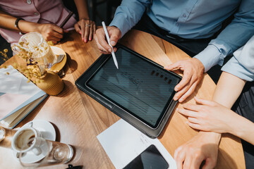 A group of colleagues gathers around a wooden table with a tablet, phones, and papers, discussing strategies, data, and ideas in a coffeehouse setting.