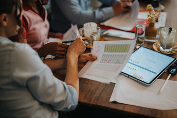 A group of professionals collaborate at a wooden table, reviewing reports and charts on paper and a tablet. They share ideas, take notes, and plan a project in a casual office setting.