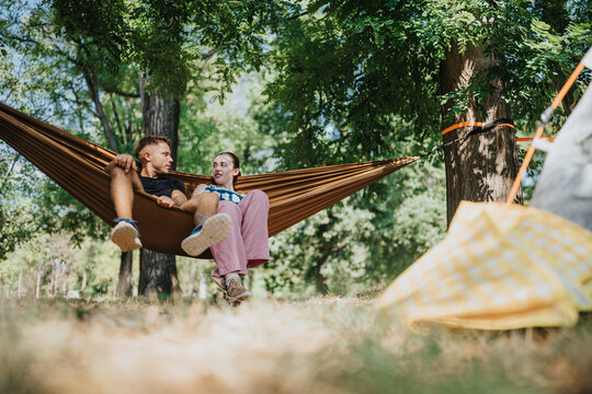 A couple lounges in a brown hammock tied between trees in a lush park. Nearby a yellow camping tent adds a cozy outdoor vibe, capturing relaxation and nature.