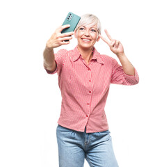 Trendy middle aged woman holding in hands smartphone and making selfie while showing v-sign, isolated on white background. Adult woman using smart phone.