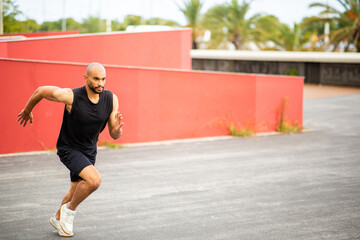Athletic man with shaved head and beard sprinting sideways outdoors on asphalt track