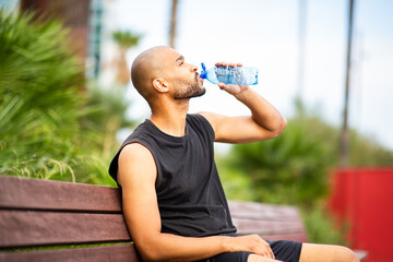 Athletic man with shaved head and beard drinking water on bench during outdoor break