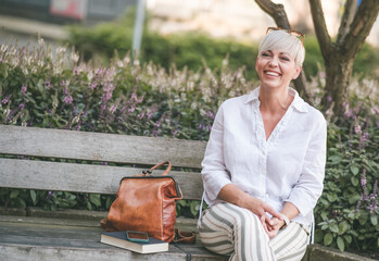 Woman with blond short hair. Cheerful female person looking to side dressed in white summer shirt while sitting on bench. Outdoors, blurred nature background.