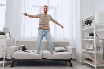 Energetic man happily dancing on the couch, surrounded by a bright, modern living room, enjoying leisure time, and embracing the joy of home life.