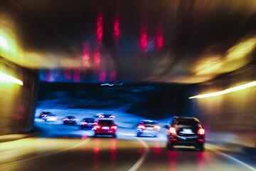 Multiple cars are captured with motion blur as they travel at speed through a brightly lit tunnel under a bridge at night. Transportation, lifestyles, or infrastructure design element.