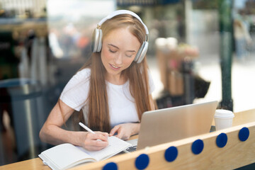 Young woman studies at a cafe with headphones and laptop in sunny outdoor setting