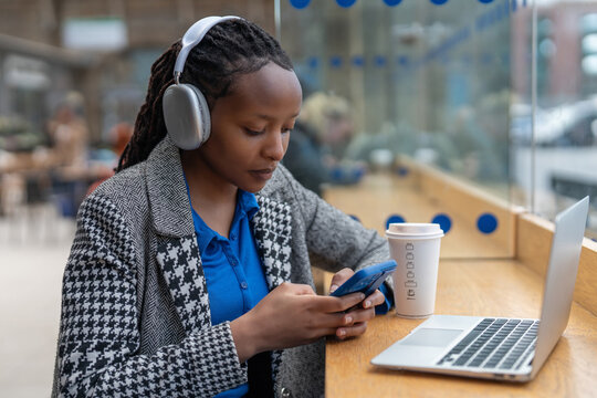 Young woman uses smartphone while listening to music and working on laptop in a busy cafe