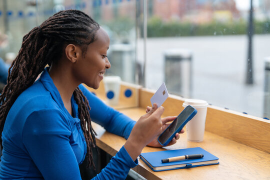 Woman uses smartphone and card while sitting at cafe table by window in urban setting
