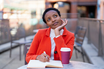 Young woman in bright orange coat thinks while holding a notebook and coffee cup outdoors