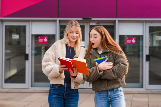 Two friends excitedly reading books outside a modern building during the day
