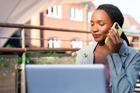 Young woman engages in a phone call while working on a laptop outdoors in a sunny urban setting
