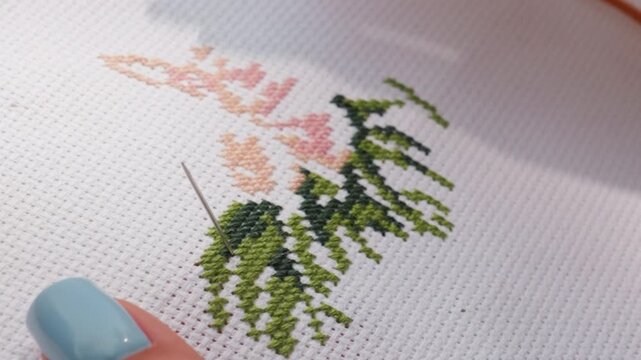 Close up designer embroidering fabric, needle passing through cross stitch pattern, blue nail visible, green and peach threads forming leafy design, precise work on white canvas under soft daylight