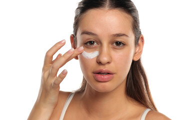 Young woman with long dark hair applying eye cream under her eye, studio shot on a white background