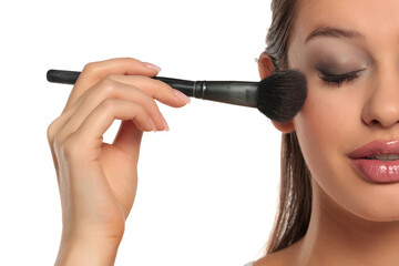 Studio shot of a woman&rsquo;s half face with eyes closed applying blush with a makeup brush on a white background