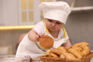Cute baby in chef uniform taking bun from basket at light marble table indoors