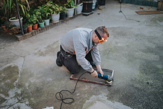 A man wearing safety goggles works with an angle grinder outdoors, kneeling on a concrete surface to sand a metal frame. Power cord lies nearby, plants line the background.