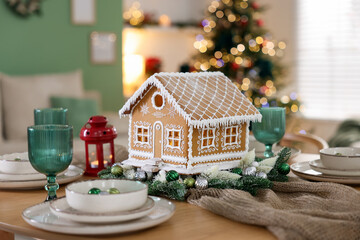 Festive table setting with beautiful gingerbread house against blurred lights indoors, closeup. Christmas atmosphere