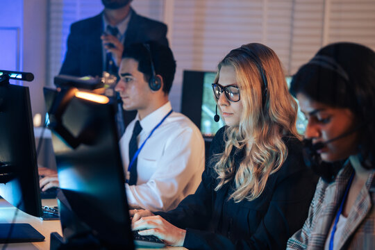 Team of dedicated customer service agents works on computers with headsets in a dark office. Technical support, IT helpdesk, call center staff, and professional late night or emergency operations