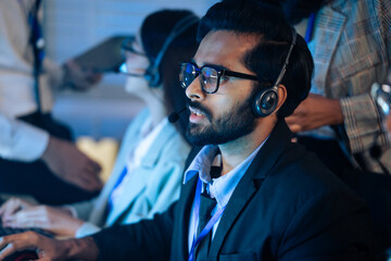 Customer service agent wears a headset talking to a client in a dark call center. Support, IT helpdesk staff, and late night corporate operations and assistance telemarketing