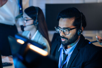 Indian man with team in a headset working call center in a control room. Customer support, IT helpdesk service, and professional telephone operations, telemarketing, sales agent at late night office