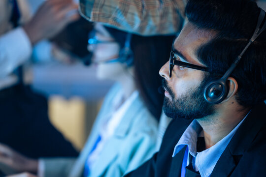 Indian customer service representative wearing headset listening to a customer in a modern call center..  Agent is providing dedicated tech support and problem-solving with diverse team at night shift