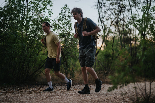 Two young men hike through dense woodland, carrying rugged backpacks and wearing casual outdoor gear. The trail is rocky and shaded, capturing a moment of fitness, teamwork, and nature exploration. - Powered by Adobe