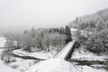 Snow blankets the landscape as a bridge spans over a gently flowing river. Trees shimmer with frost, creating a peaceful winter atmosphere in the hidden countryside