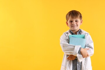 Smiling little boy in laboratory coat with books on yellow background, space for text. Child and...