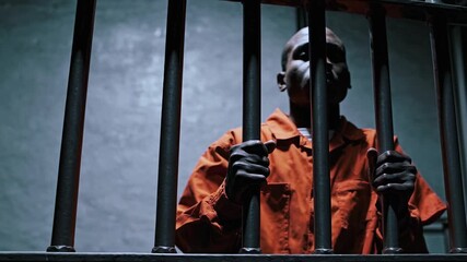 Prisoner behind bars in dim jail cell. Person in orange inmate clothing standing behind prison bars in dim detention cell. Confinement and criminal justice. Incarceration, justice system concept - Powered by Adobe