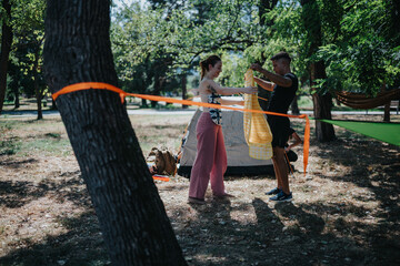Two friends share a moment while pitching a hammock near a tent in a shaded park, enjoying outdoor...