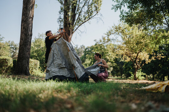Two people pitch a silver tent in a lush park under clear skies. A relaxed outdoor camping scene that emphasizes teamwork, nature, and weekend adventure.