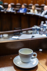 An empty coffee cup on a wooden desk with the blurred background of a plenary session in a government assembly, symbolizing a break or the long hours of political debate and decision-making.
