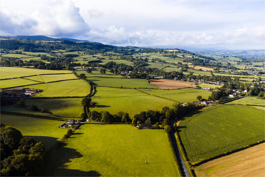 Aerial view of lush green fields contrasting with dark tree lines and distant rolling hills under a dynamic sky, St. Davids, Wales, United Kingdom.