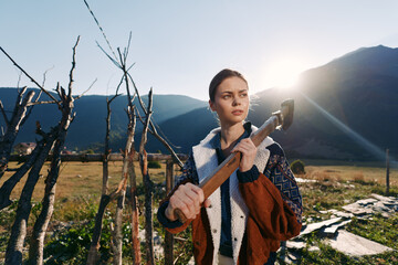 Fototapeta premium Woman with axe near a wooden fence in rugged mountains on a small farm at sunrise, rural portrait capturing determination and rustic outdoor lifestyle with sunlight on meadow.