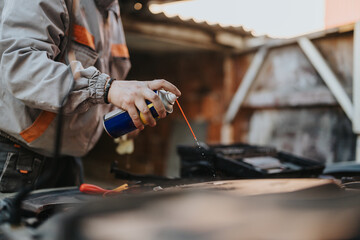 A technician wearing a work jacket sprays a liquid lubricant over the engine bay in a garage setting. The scene captures auto maintenance, repair work, and hands-on car care.
