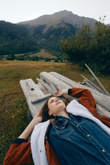 Fototapeta premium woman outdoors in meadow and mountains lying on wood planks, portrait of relaxation under open sky, casual denim jacket and cozy coat in peaceful natural landscape