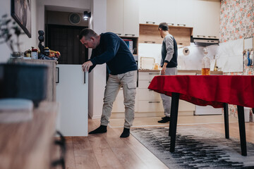 Two men in a bright kitchen move and organize a cabinet as a table with a red cloth stands nearby,...