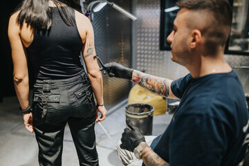 A tattoo artist in gloves inks a design on a woman's forearm in a contemporary studio, with metallic walls and equipment in the background.