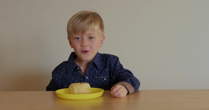 A blond boy in a blue shirt is smelling a donut, also known as a French cruller. He is smiling and showing that he likes this food.