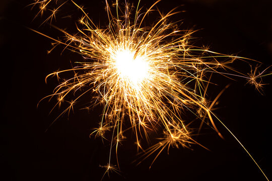 A close-up shot of a bright golden sparkler exploding with sparks against a deep black background. Symbolizes celebration, joy, and New Year's fireworks.