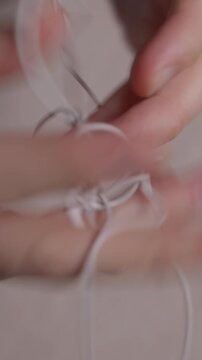 Vertical close up of a mans hands trying to untangle white wired earphones. The knots and small movements highlight a familiar everyday struggle with personal tech accessories. High quality 4k