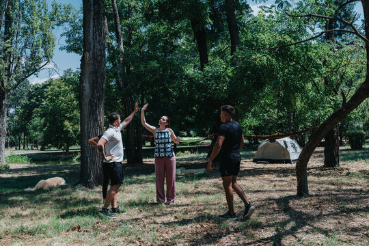 A small group of friends share a moment in a forested park, with a tent in the background. They stand and gesture, creating a relaxed outdoor camping scene.
