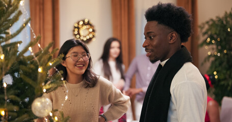 During a group dinner meeting, friends are talking near a Christmas tree. A woman with glasses and a man with dark hair and stubble tell each other about decorations and baubles.