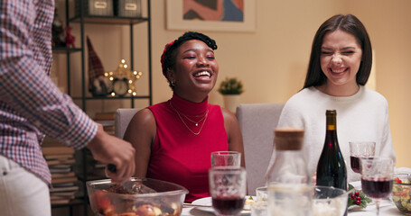 Fototapeta premium During the dinner, guy in the shirt brought main dish, cuts the meat and puts it on guests' plates. Girl in red clothes is waiting for portion. Girls are laughing and chatting.