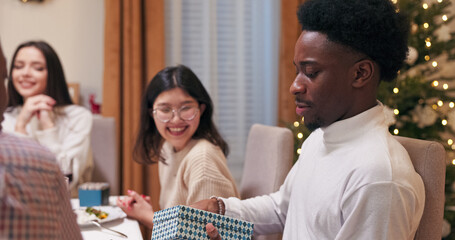 Friends are smiling at the festive table as gifts are exchanged. A multi-ethnic guy in a white sweater will receive a gift, untie the ribbon, open the box, be happy, and show the surprise to all his