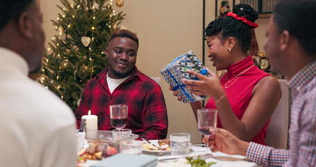 The Christmas tree flickers behind a girl in a red dress who gives a Christmas present to a boy in a red shirt. Friends applaud, smile, and rejoice as the student opens the box and is pleasantly